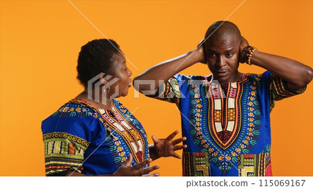 Furious girlfriend shouting at her boyfriend and fighting, having disagreement over orange background. Frustrated man covering his ears and trying to ignore his wife yelling in studio. Furious girlfriend shouting at her boyfriend and fighting, having disagreement over orange background. Frustrated man covering his ears and trying to ignore his wife yelling in studio. 115069167