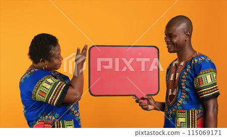 Married ethnic people pointing at red speech bubble icon made from cardboard for web promotions and advertisements. Young man and woman promoting something with isolated billboard sign. 115069171