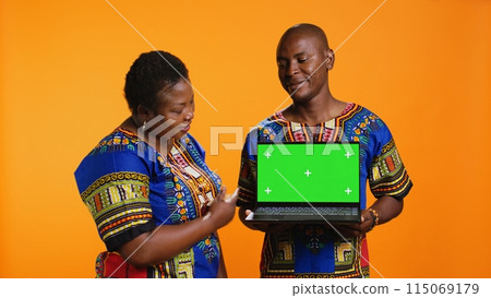 African american partners holding laptop with greenscreen on camera, presenting isolated display with chromakey and standing over orange background. Man and woman showing blank mockup. 115069179