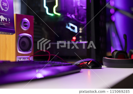 Neon lit components on opened gaming PC device next to keyboard, speakers, wired mouse and headphones in empty apartment. Computer in home with colorful gpu and cpu parts, close up shot 115069285