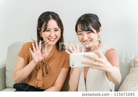 Asian woman sitting on a sofa and waving while making a video call on her smartphone 115071095