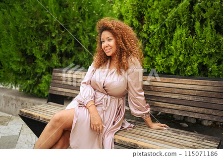 Young beautiful African American woman enjoys a walk in the summer public green park. A happy woman plus size wearing silk pink dress smiles walks through the city streets, resting on a bench. 115071186