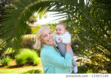 Beautiful happy smiling senior elderly woman holding on hands cute little baby boy. Grandmother and grandson having fun time together at tropical summer day. Multigenerational family concept. 115071189