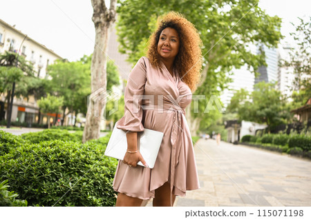 Attractive happy stylish plus size African American woman student freelancer afro hair with laptop computer outside, big urban city street at summer. Diversity. Remote office work, distance education. Attractive happy stylish plus size African American woman student freelancer afro hair with laptop computer outside, big urban city street at summer. Diversity. Remote office work, distance education. 115071198