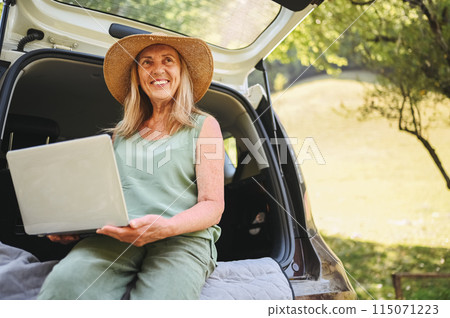 Senior happy woman working with laptop from the car trunk or boot in camper sunny summer park - digital nomad freedom lifestyle concept, travel freelancer and vacations of retired old age people.. Senior happy woman working with laptop from the car trunk or boot in camper sunny summer park - digital nomad freedom lifestyle concept, travel freelancer and vacations of retired old age people.. 115071223