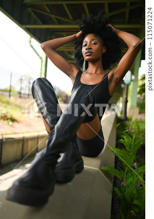 Fashion outdoor street style portrait Beautiful young African American woman posing outside on urban city landscape summer day wearing total black high leather boots 115071224