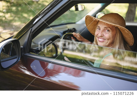 Happy senior woman driver in straw hat driving sitting in new car, smiling looking at camera enjoying journey. Driving courses and life insurance. Retired people activity and road trip concept. 115071226