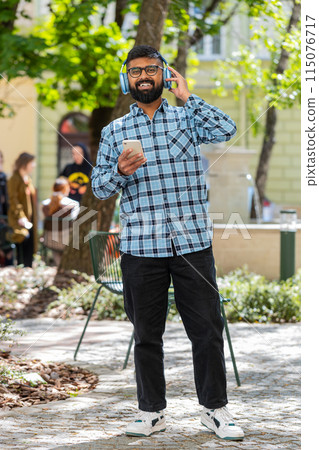 Happy Indian man dancing in wireless headphones listening favorite energetic music on city street Happy Indian man dancing in wireless headphones listening favorite energetic music on city street 115076717