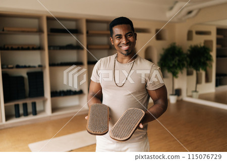 Medium shot of friendly African man holding in hands wooden Sadhu Board with nails standing in cozy living room, preparing to practice, smiling looking at camera. Concept of mindfulness, tranquility. Medium shot of friendly African man holding in hands wooden Sadhu Board with nails standing in cozy living room, preparing to practice, smiling looking at camera. Concept of mindfulness, tranquility. 115076729