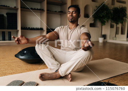 Portrait of mindful African American man meditating in lotus pose at home sitting on yoga mat with closed eyes near wooden sadhu board and metal hang drum. Concept of tranquility, mental health. Portrait of mindful African American man meditating in lotus pose at home sitting on yoga mat with closed eyes near wooden sadhu board and metal hang drum. Concept of tranquility, mental health. 115076760