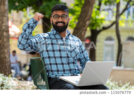 Indian happy man working on laptop celebrate success win money sitting on urban street in city Indian happy man working on laptop celebrate success win money sitting on urban street in city 115076779