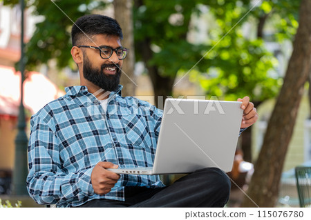 Indian man freelancer working online distant job with laptop browsing website sitting on city street 115076780