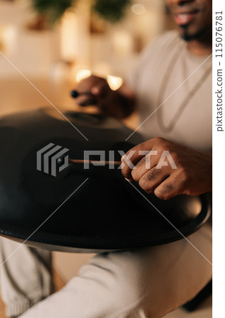 Vertical cropped shot of unrecognizable calm African man playing traditional ethnic music on glucophone using sticks sitting on floor in lotus pose. Silence calm filling room by sounds of music. 115076781