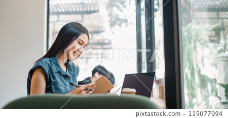 A woman is sitting at a table with a laptop and a notebook. She is smiling and she is enjoying her time 115077994