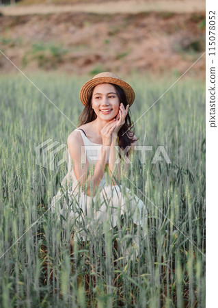 A woman is sitting in a field of tall grass, wearing a straw hat and a white dress. She is smiling and she is enjoying the outdoors 115078052