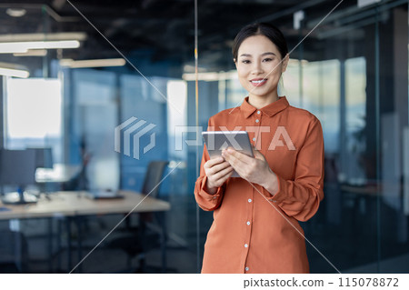 Portrait of a successful Asian female programmer with a tablet computer in her hands, an employee of a development company is smiling and looking at the camera near the window. 115078872
