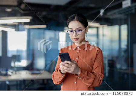 Asian woman using phone, serious thoughtful business woman inside office typing text message and browsing social media, female programmer software developer testing app online. Asian woman using phone, serious thoughtful business woman inside office typing text message and browsing social media, female programmer software developer testing app online. 115078873