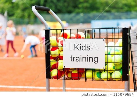 Basket cart filled colorful tennis balls sign plate Kids-training clay tennis court children playing training background. Summer sport camp. Sports youth school. Healthy child kid active lifestyle 115079694