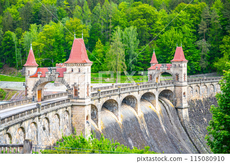 A view of the Les Kralovstvi Dam in Czechia, showcasing the intricate archway and stonework of the structure. The dam stands tall and imposing, surrounded by lush greenery. 115080910