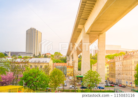 Warm sunlight bathes the Nusle Bridge and surrounding streetscape in Prague, highlighting the contrast between the bridges modern structure and the classic city architecture. Prague, Czechia 115083116