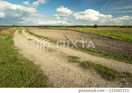 Gravel road next to a plowed field and clouds on the sky Gravel road next to a plowed field and clouds on the sky 115085353