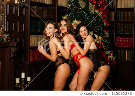 Three seductive females posing in lingerie under fir tree. Studio shot. Christmas content. Three seductive females posing in lingerie under fir tree. Studio shot. Christmas content. 115085378
