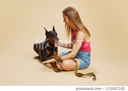 Girl in pink top and denim shorts sits on floor with her Doberman, holding pet close and smiling against beige studio background. Girl in pink top and denim shorts sits on floor with her Doberman, holding pet close and smiling against beige studio background. 115087185