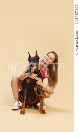 Girl with sits and hugs from behind her lovely purebred Doberman dog and cheerful smiling against beige studio background. Girl with sits and hugs from behind her lovely purebred Doberman dog and cheerful smiling against beige studio background. 115087196