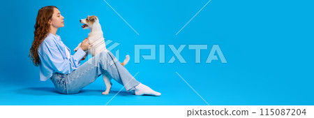 Banner. Girl with curly hair plays with her dog, seated on floor against vibrant blue studio background with copy space. 115087204