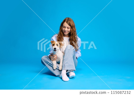 girl with long curly hair sits on the floor holding a small dog on her lap and smiling against a bright blue background. 115087232