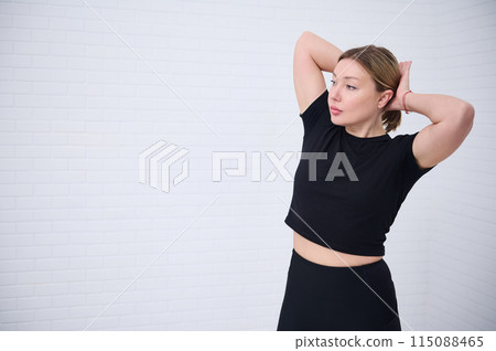 Beautiful Caucasian athletic woman 30s in black sportswear, tying a ponytail, confidently looking way, isolated over white wall background with copy advertising space 115088465