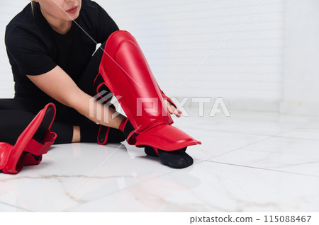 Selective focus on legs of a woman boxer fighter putting on red kickboxing leg pads guard. Sparring shin guards. Martial art, combat, challenge. Kickboxing concept Selective focus on legs of a woman boxer fighter putting on red kickboxing leg pads guard. Sparring shin guards. Martial art, combat, challenge. Kickboxing concept 115088467