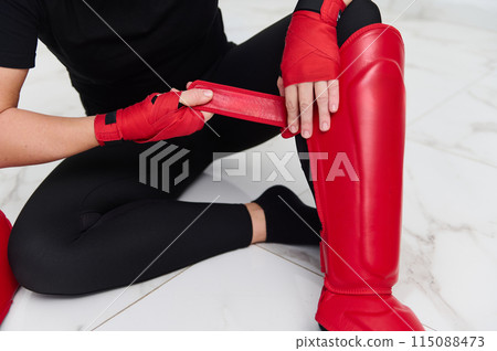 Selective focus on legs of a woman boxer fighter putting on red kickboxing leg pads guard. Sparring shin guards. Martial art, combat, challenge. Kickboxing concept. Selective focus on legs of a woman boxer fighter putting on red kickboxing leg pads guard. Sparring shin guards. Martial art, combat, challenge. Kickboxing concept. 115088473