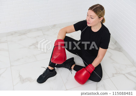 View from above of a blonde female boxer fighter wearing red boxing gloves, getting ready for combat, sitting over white wall background 115088481