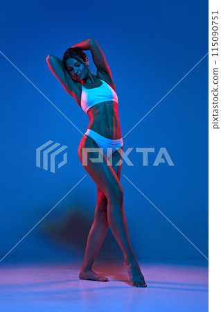 Full length portrait of young attractive woman posing raising hands showing her fit forms in neon light against gradient blue background. Full length portrait of young attractive woman posing raising hands showing her fit forms in neon light against gradient blue background. 115090751