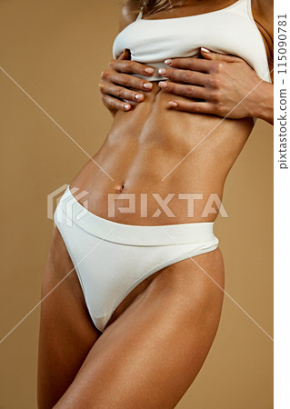 Cropped photo of woman's toned midsection in white underwear, with her hands on her chest against beige studio background. 115090781