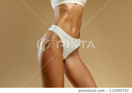 Detailed view of woman's sculpted abs and hips in white lingerie against beige studio background. Model with fit figure. 115090782