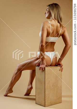 Rear view of toned woman in white lingerie sits gracefully on wooden block, emphasizing her fit against beige studio background. Rear view of toned woman in white lingerie sits gracefully on wooden block, emphasizing her fit against beige studio background. 115090794