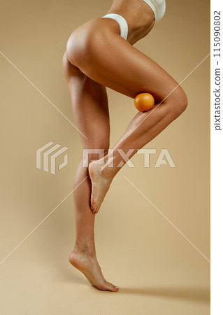 Cropped photo of woman's fit legs and buttocks, holding orange between her knee and thigh against beige studio background. Cropped photo of woman's fit legs and buttocks, holding orange between her knee and thigh against beige studio background. 115090802