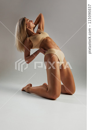 Fit woman with long blonde hair poses on her knees, arching her back and stretching in beige lingerie against grey studio background. Fit woman with long blonde hair poses on her knees, arching her back and stretching in beige lingerie against grey studio background. 115090837