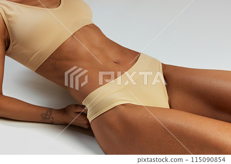 Close-up of woman's toned midsection, highlighting her defined abs and beige ribbed lingerie against grey studio background. Close-up of woman's toned midsection, highlighting her defined abs and beige ribbed lingerie against grey studio background. 115090854