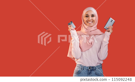 Mobile banking. Online shopping. Satisfied cheerful happy woman in hijab showing credit card phone isolated on red empty space background. Mobile banking. Online shopping. Satisfied cheerful happy woman in hijab showing credit card phone isolated on red empty space background. 115092248