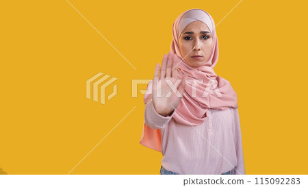 Enough gesture. Female protest. Concerned dissatisfied frowning woman in hijab warning with outstretched hand palm isolated on orange empty space background. Enough gesture. Female protest. Concerned dissatisfied frowning woman in hijab warning with outstretched hand palm isolated on orange empty space background. 115092283