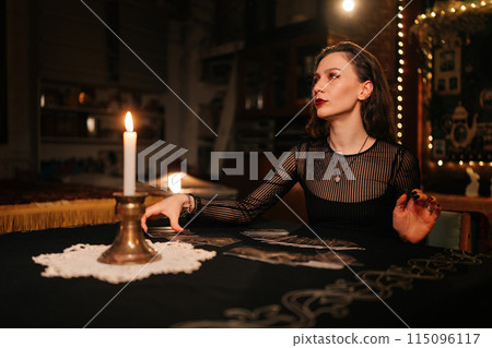 Portrait of enigmatic fortuneteller laying out tarot cards, reading signs symbols on magic cards and talks with client sitting at table in dark esoteric room with mystical atmosphere. 115096117