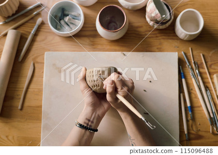 From above view of hands of unrecognizable artisan using clay carving tool to decorate ceramic bowl 115096488