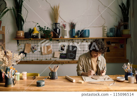 Cheerful female artisan sitting at wooden table in pottery workshop creating leaf-shaped plate, copy space 115096514