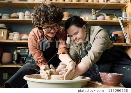 Low angle view shot of two diverse women wearing aprons having fun during pottery class shaping clay on wheel together Low angle view shot of two diverse women wearing aprons having fun during pottery class shaping clay on wheel together 115096568