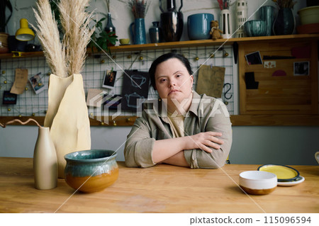 Medium portrait of young Caucasian woman with disability sitting at wooden table in pottery workshop looking at camera Medium portrait of young Caucasian woman with disability sitting at wooden table in pottery workshop looking at camera 115096594