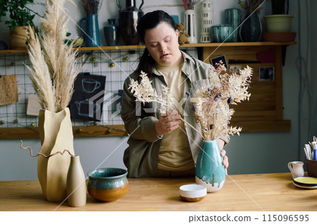 Creative young woman with Down syndrome standing at table in workshop putting fried flowers into ceramic vase Creative young woman with Down syndrome standing at table in workshop putting fried flowers into ceramic vase 115096595