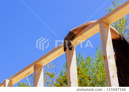 As framer installs beams using an air nail hammer, he nails wooden planks to frame As framer installs beams using an air nail hammer, he nails wooden planks to frame 115097166